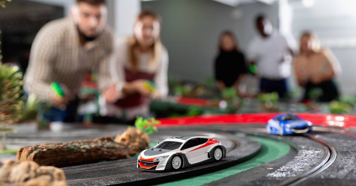  Two slot cars, one red and white and the other blue, race down a large track while people in the background control them.