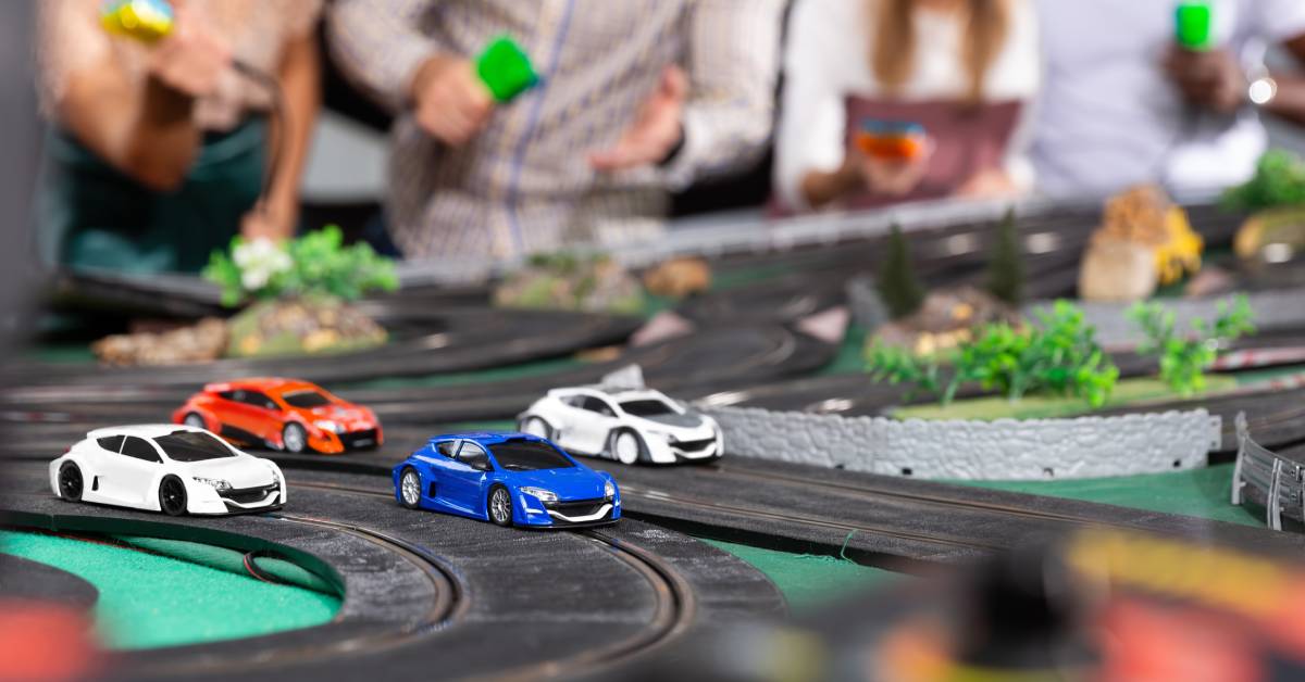 A group of people with controllers gathered around a slot racing track, watching cars speed across the layout.